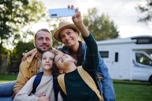 Uma família jovem e feliz com duas crianças tirando selfie com caravana ao fundo ao ar livre.