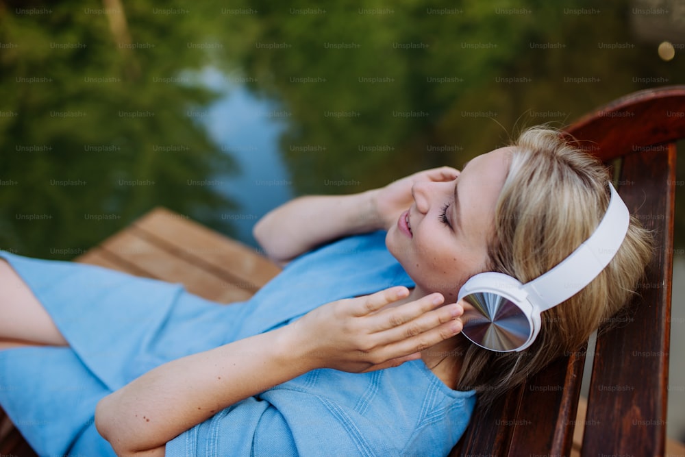 Una mujer relajada con auriculares escuchando música tumbada en un muelle junto a un lago natural en verano