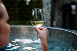 An unrecognizable young woman relaxing with glass of wine in hot tub outdoor in nature.