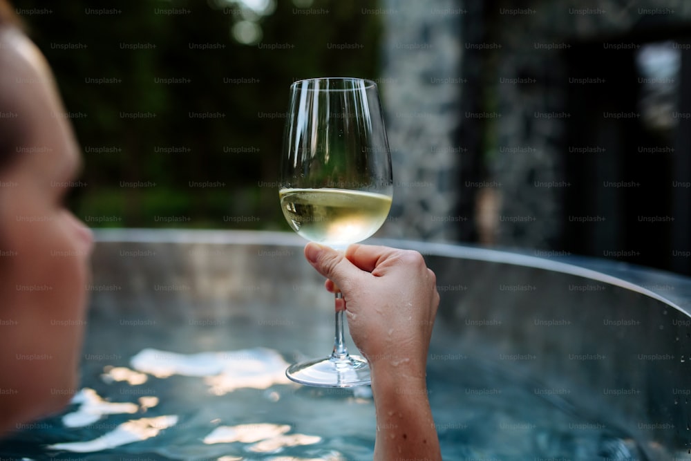 Una joven irreconocible relajándose con una copa de vino en una bañera de hidromasaje al aire libre en la naturaleza.