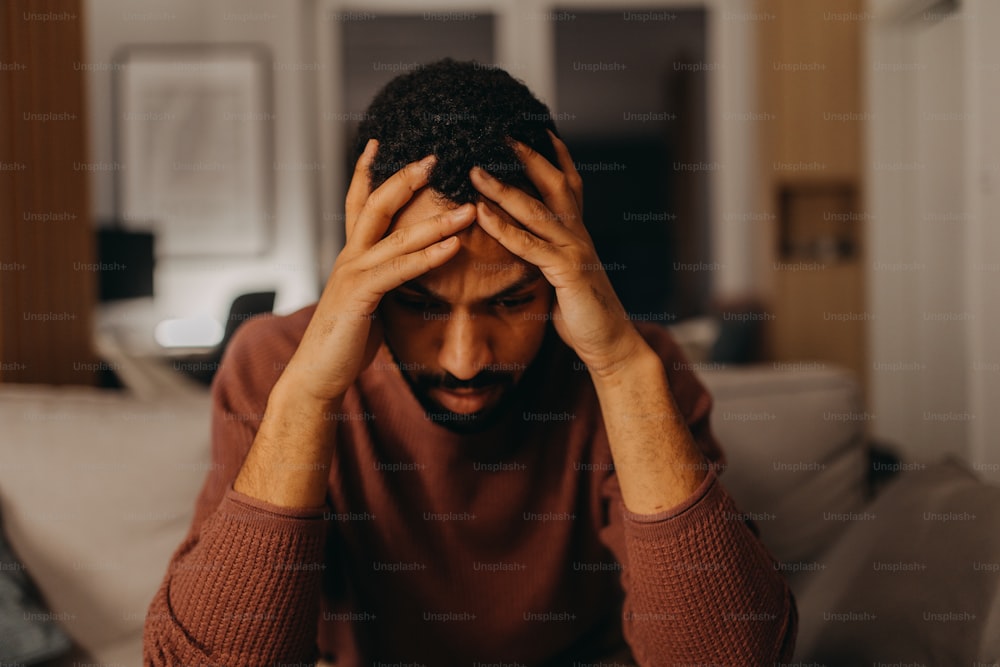 Ein Porträt eines depressiven jungen afroamerikanischen Mannes mit dem Kopf in den Händen, der zu Hause sitzt.