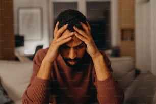 Um retrato od deprimido jovem afro-americano homem com a cabeça nas mãos sentado em casa.