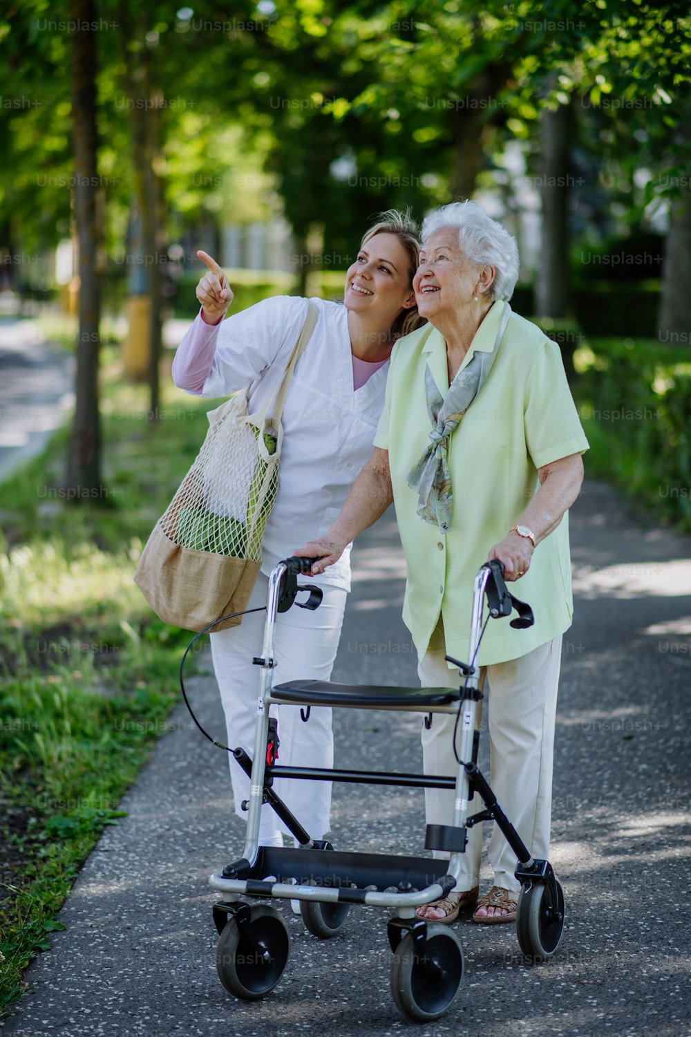 買い物袋を持って公園を歩行器で散歩する年配の女性と介護者。