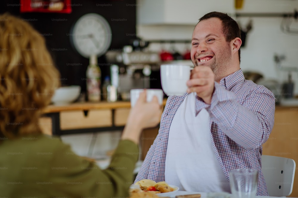 A portrait of happy man with Down syndrome with his mother at home having breakfast together.