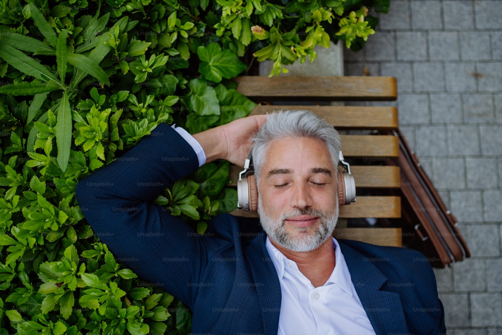 A top view of mature businessman enjoying music in headphones relaxing on bench in city park during break at work.