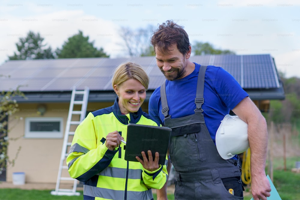 A man and woman solar installers engineers with tablet while installing solar panel system on house.