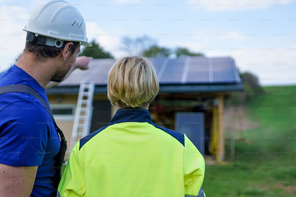 Un hombre y una mujer ingenieros instaladores solares con tableta mientras instalan el sistema de paneles solares en la casa, vista trasera.