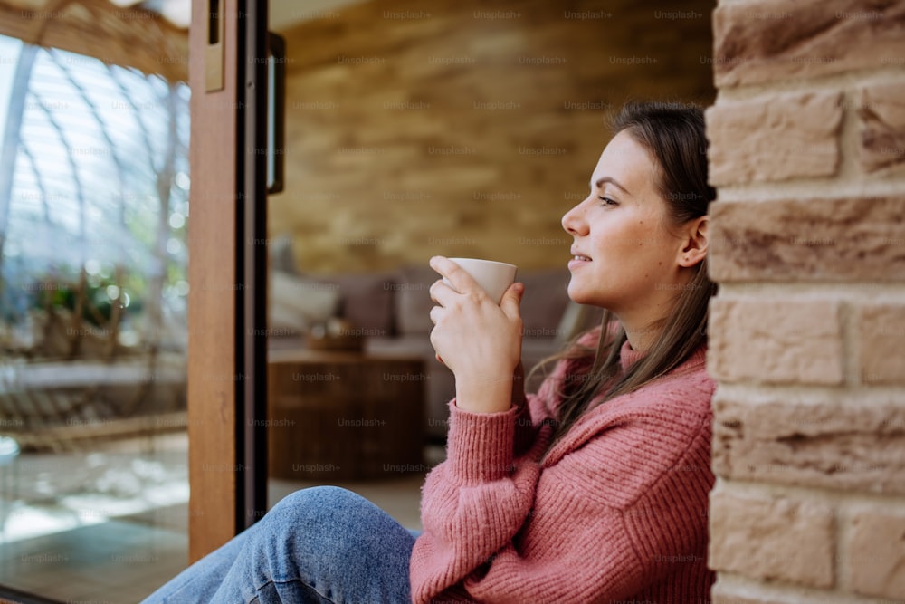 Uma jovem sorridente com rosto sonhador bebendo café da manhã e sentada no pátio de sua casa