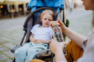 An unrecognizable mother holding zero waste water bottle and opening it in front of her little child in stroller, sustainable lifestyle.
