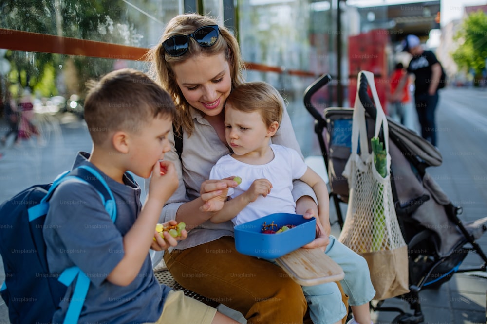 어린 아이들과 함께 도시의 버스 정류장에서 기다리고 과일 간식을 먹는 젊은 어머니.