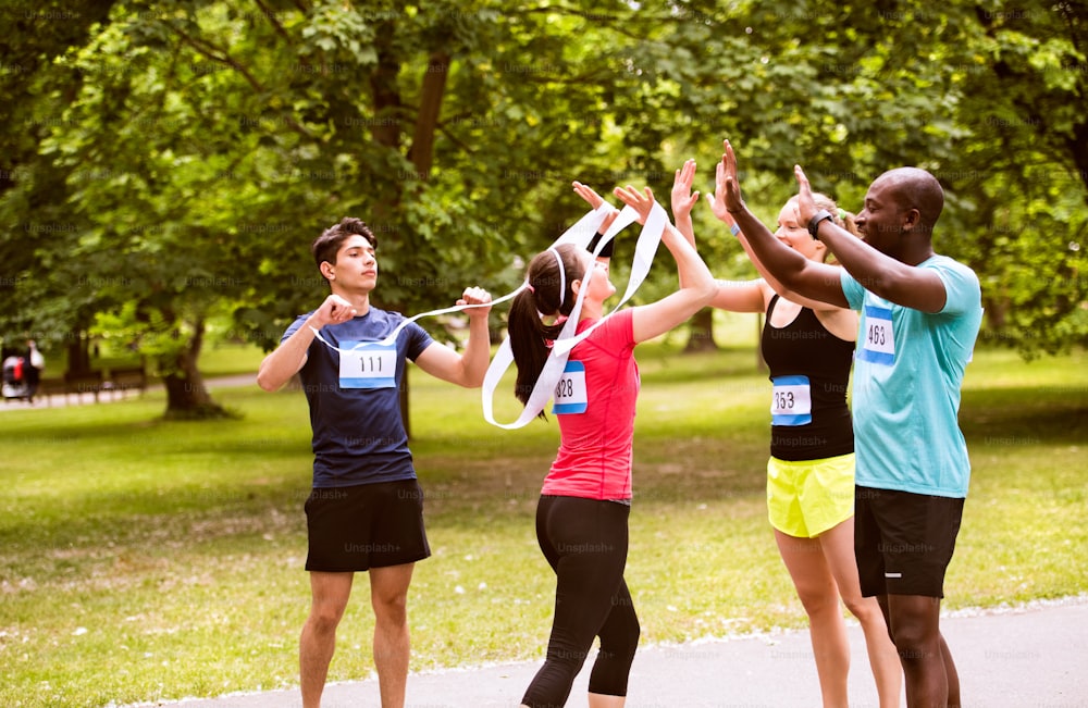 Group of young fit friends at the finish line happy after completing race, giving high five to each other. Green sunny park.