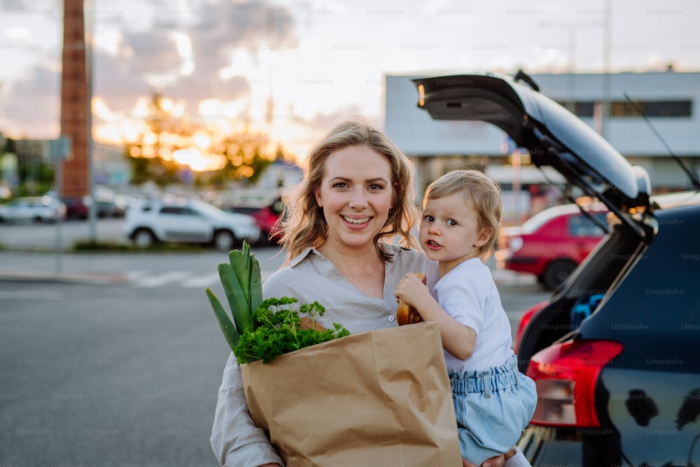 Una madre joven con una hija pequeña después de ir de compras sosteniendo bolsas de compras de residuo cero con comestibles cerca del automóvil.