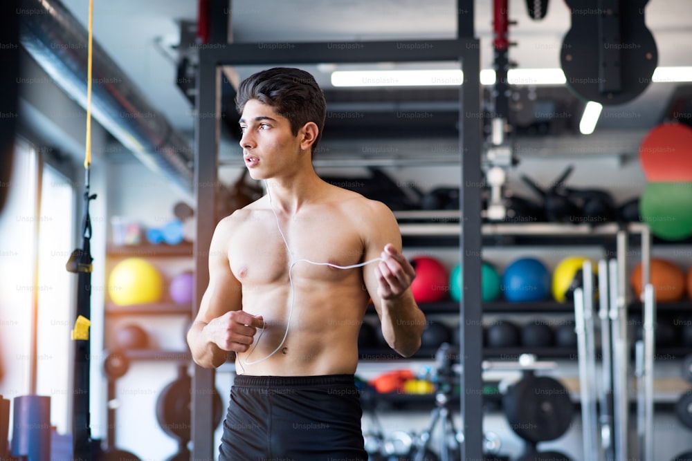 Joven hispano guapo hombre de fitness en el gimnasio sosteniendo auriculares, escuchando música