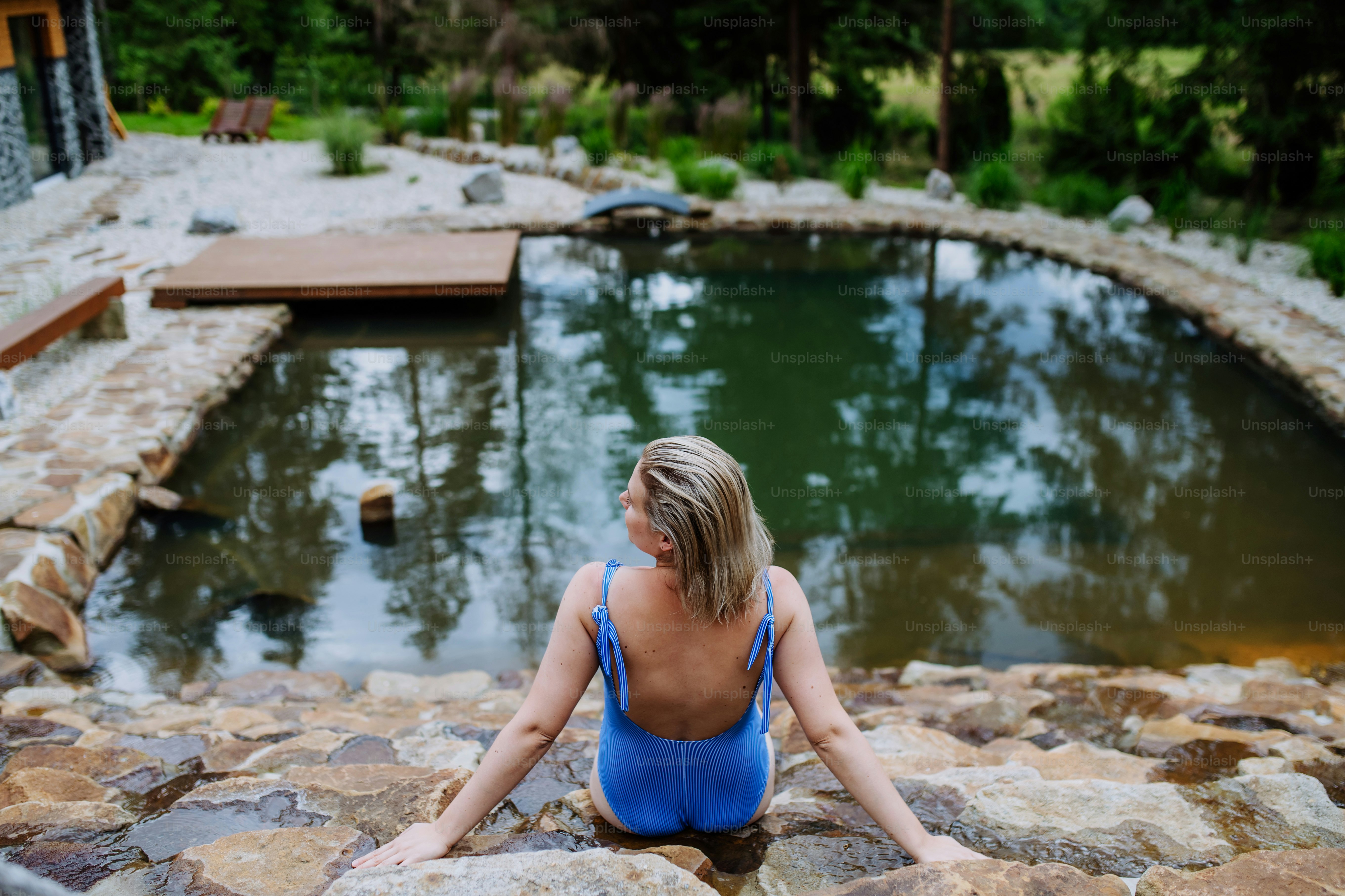 Una vista posteriore di una giovane donna in costume da bagno seduta vicino allo stagno naturale del cortile durante le vacanze estive in montagna.