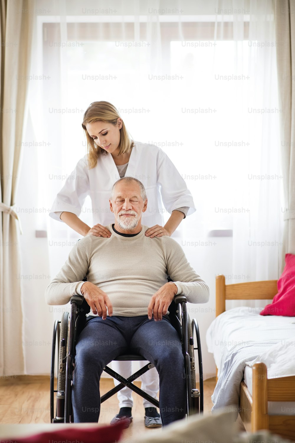 Health visitor and a senior man in a wheelchair during home visit. A nurse or a physiotherapist giving man shoulder massage.