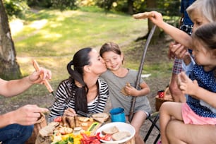 Beautiful family enjoying camping holiday in forest, eating together. Barbecue with drinks and food.