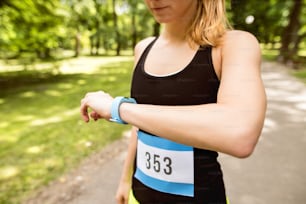 Unrecognizable young woman setting up watch before going for a run in green sunny summer park.