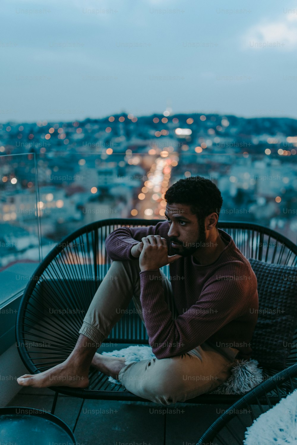 Ein Porträt eines depressiven jungen afroamerikanischen Mannes, der abends auf einem Balkon mit Blick auf die Stadt sitzt.