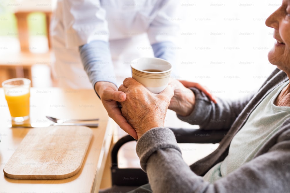 Visitador de salud irreconocible y una mujer mayor durante la visita domiciliaria. Una enfermera dando té a una anciana sentada a la mesa. Cerrar.