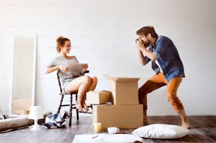 Young married couple moving in new house, woman with tablet sitting on chair, man taking pictures of her.
