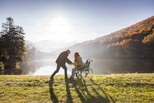 Coppia anziana attiva durante una passeggiata in una splendida natura autunnale. Un uomo e una donna su una sedia a rotelle in riva al lago al mattino presto.