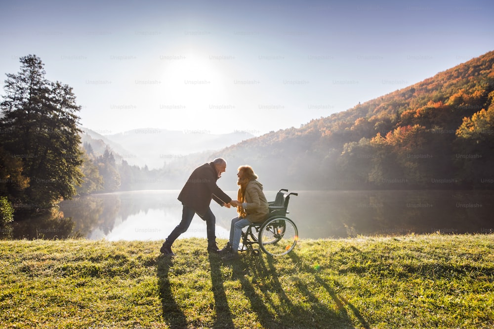 Aktives Seniorenpaar bei einem Spaziergang in einer wunderschönen Herbstnatur. Ein Mann und eine Frau im Rollstuhl am See am frühen Morgen.