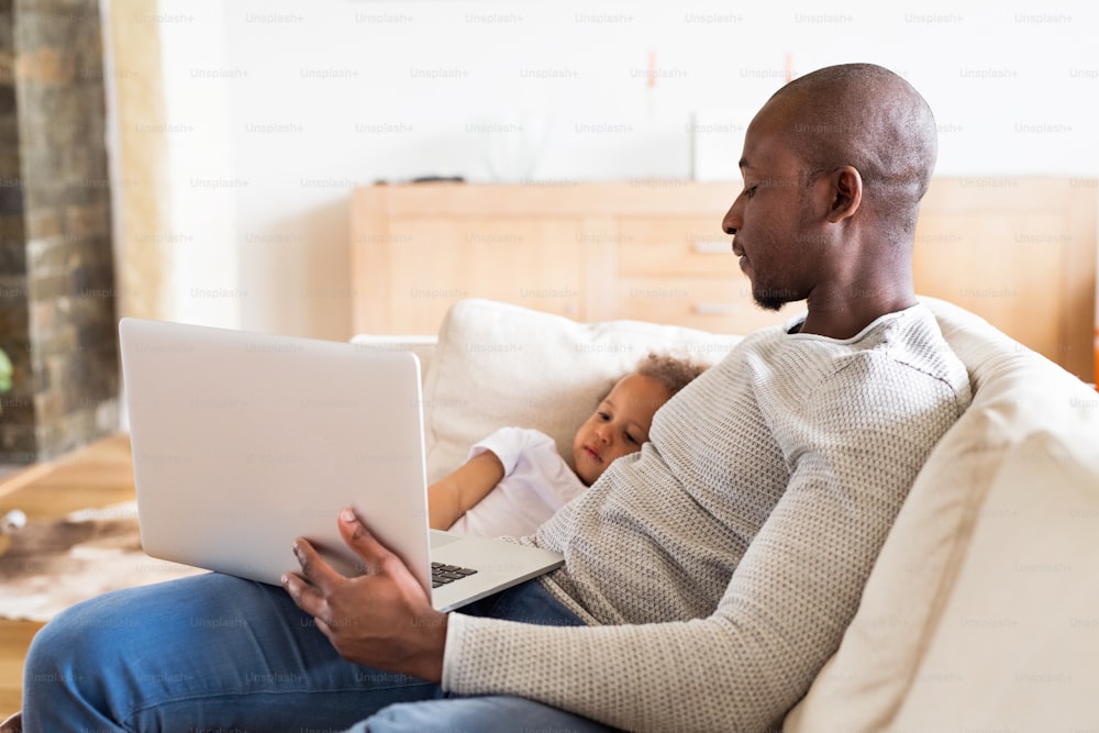 Young afro-american father at home with his cute little daughter sitting on couch watching something on laptop.