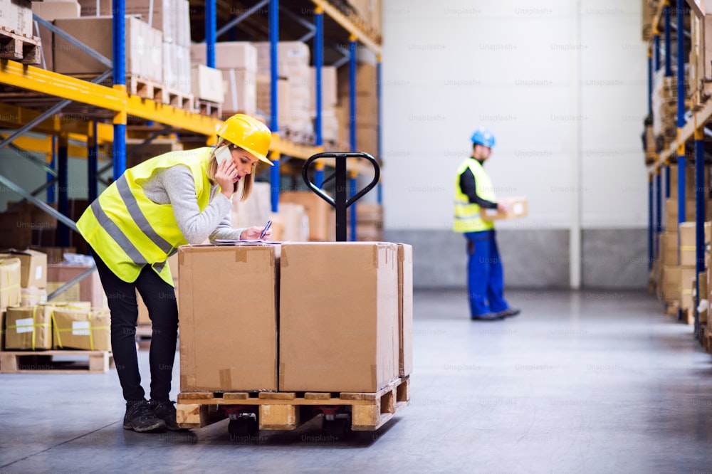 Young woman warehouse worker with smartphone, making a phone call. A man worker holding a box.