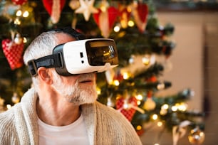Senior man in front of illuminated Christmas tree inside the house trying on VR glasses. Close up.