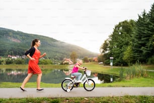 Young beautiful mother with her cute little daughter in nature by the lake. Daughter is cycling and smilling, mother is running.