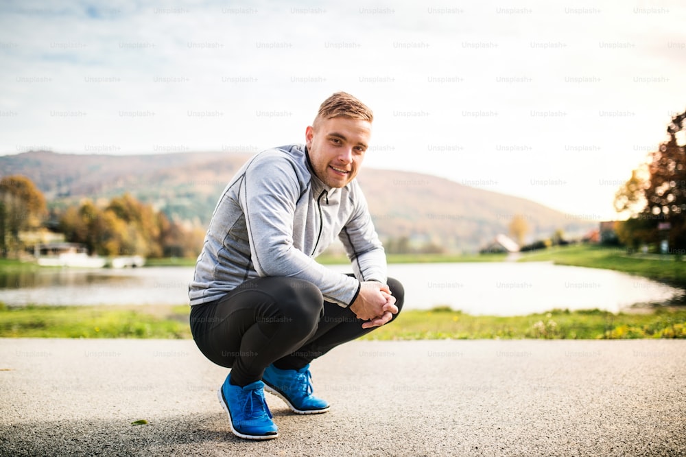 Handsome young hipster runner in gray jacket outside in colorful sunny autumn nature, squatting down on an asphalt path.