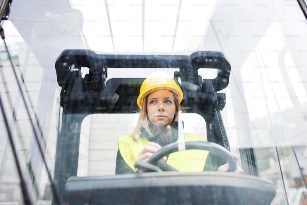 Female forklift truck driver in an industrial area. A woman sitting in the fork lift outside a warehouse. Shot through glass.