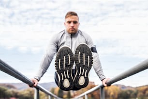 Young man with smartphone in gray jacket exercising outside in autumn nature. Parallel bars L-sit.