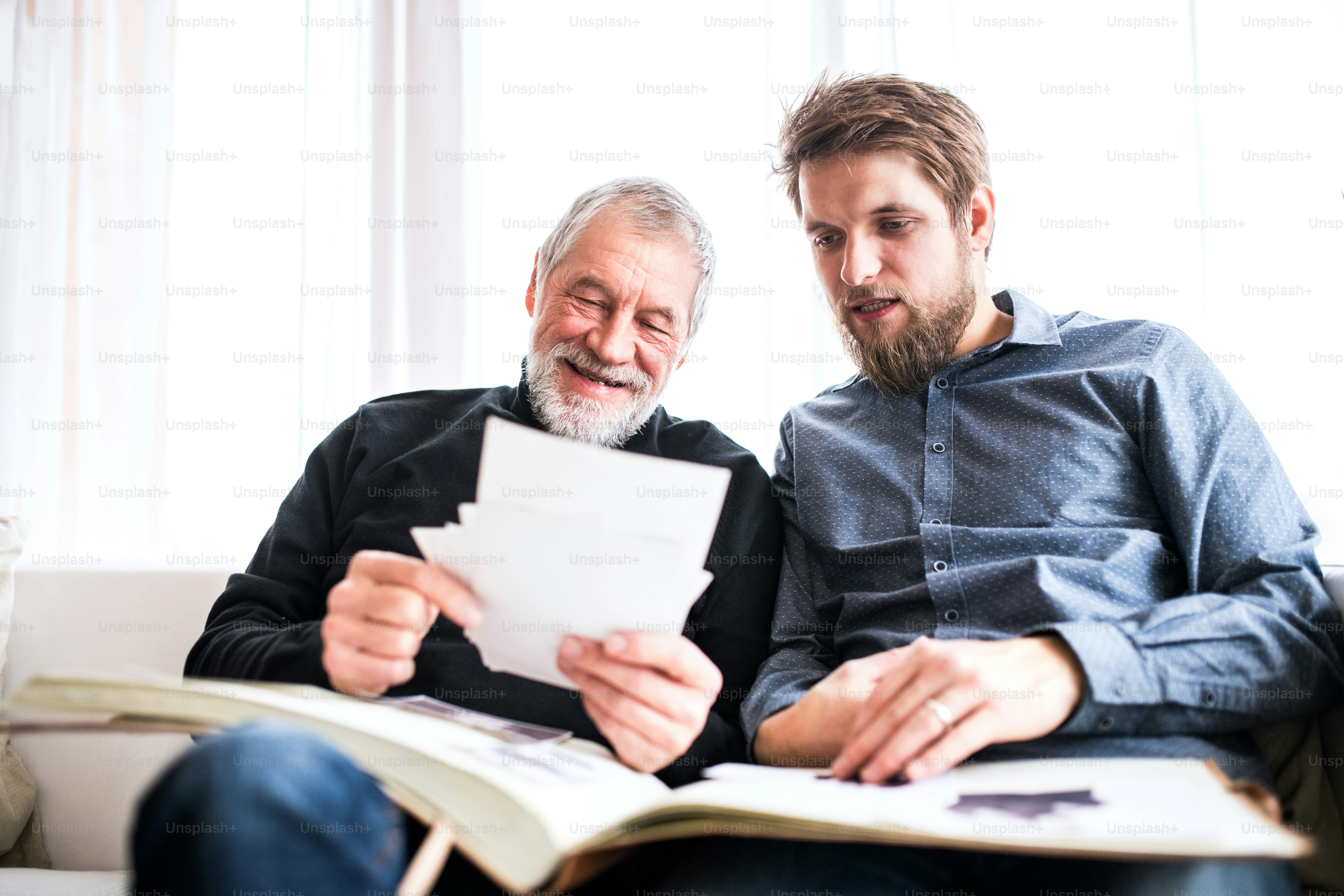 Hipster son and his senior father at home, looking at old photographs. Two generations indoors.