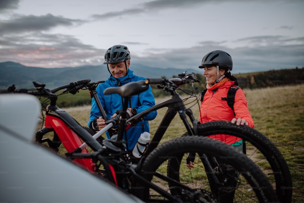 An active senior couple loading their bicycles on car carrier outdoors in nature in autumn day.