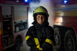 Eine mittelerwachsene schmutzige Feuerwehrfrau, die nachts mit einem Feuerwehrauto im Hintergrund in die Kamera schaut.