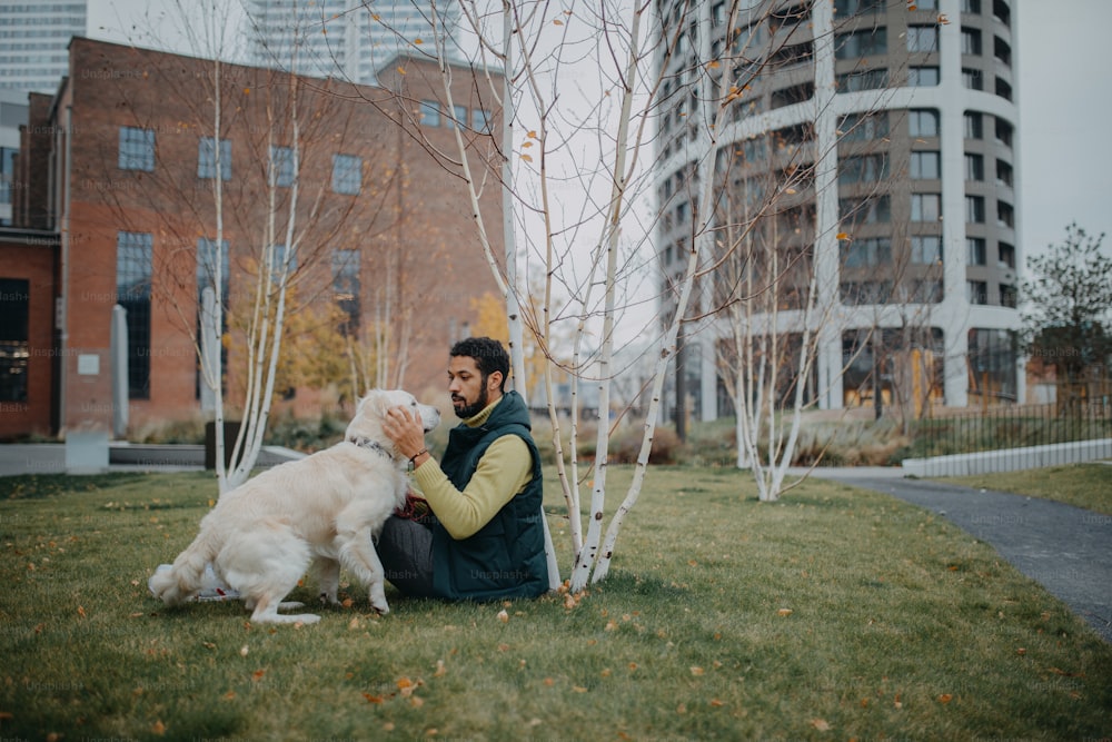 Un jeune homme heureux assis sur l’herbe avec son chien à l’extérieur en ville.