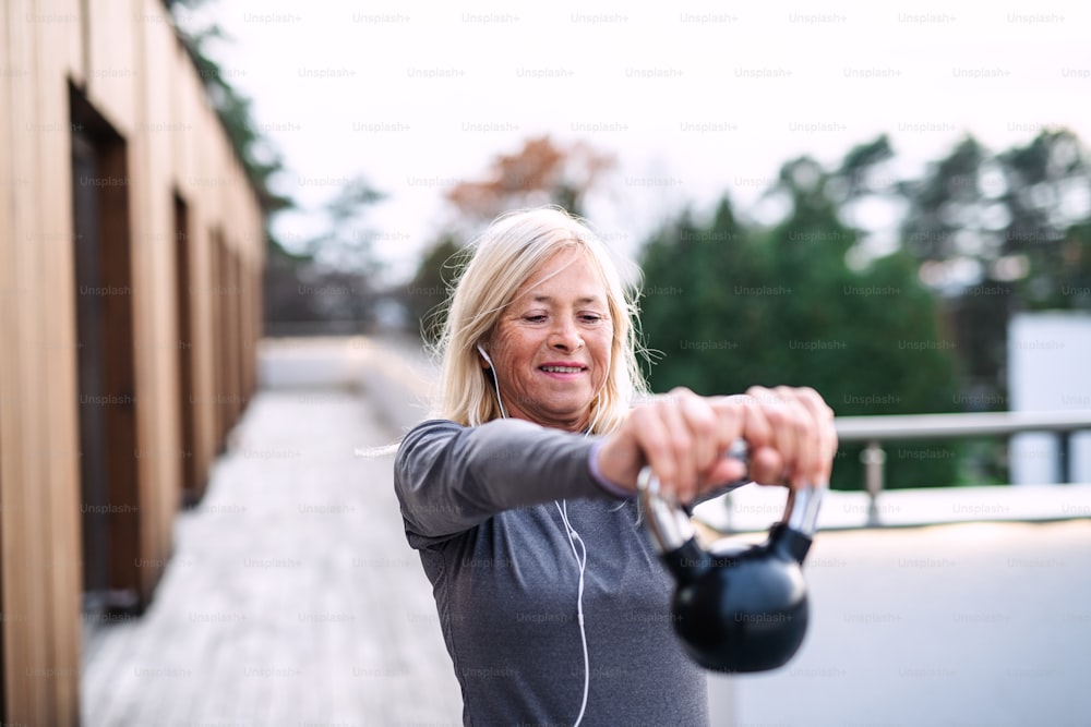 A senior woman with earphones and kettlebell outdoors doing exercise on terrace.