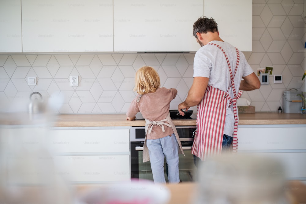 A rear view of small boy helping father indoors in kitchen with making pancakes.