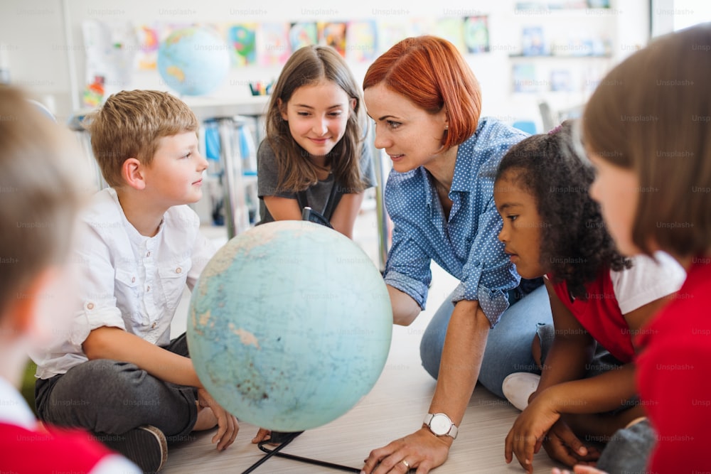 A group of small school kids with unrecognizable teacher sitting on the floor in class, learning geography.