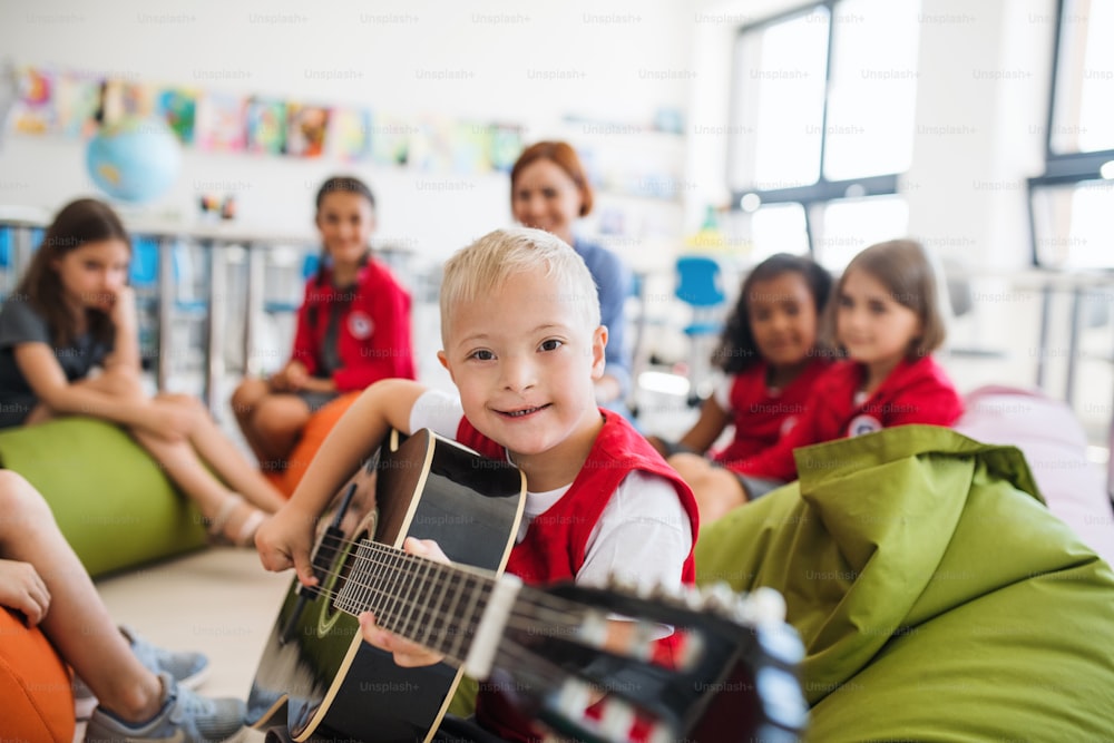 Um menino com síndrome de down com crianças da escola e professor sentado no chão em sala de aula, tocando guitarra.