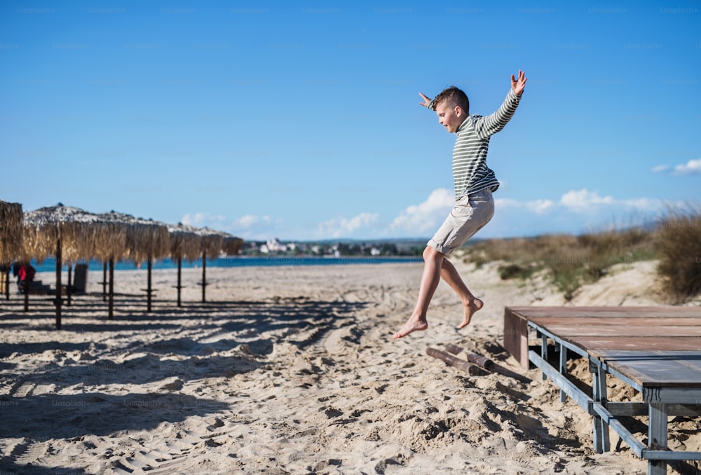 쾌활한 작은 소년이 모래 해변에서 야외에서 놀고 점프합니다.
