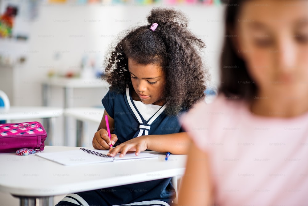 Ein kleines, gemischtrassiges glückliches Schulmädchen sitzt am Schreibtisch im Klassenzimmer und schreibt.