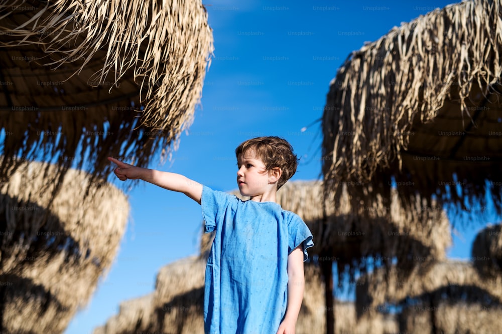 A small girl standing among straw parasols outdoors on beach.