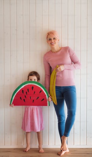 A young woman with small daughter standing indoors at home, holding toy fruit.