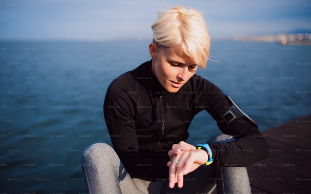 A midsection of young sportswoman sitting outdoors on beach, using smartwatch.