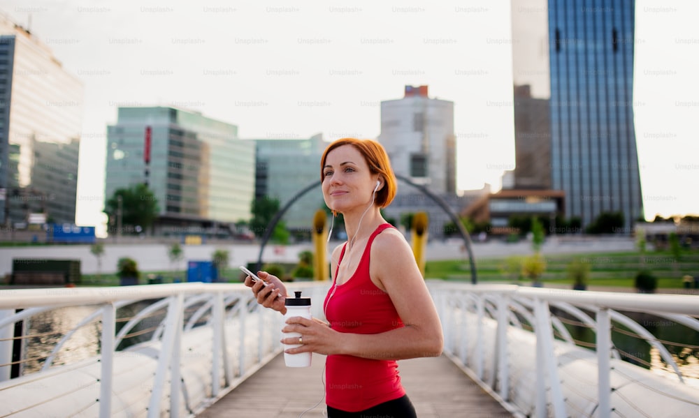 Young woman runner with earphones in city, using smartphone when resting.