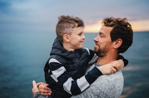 A portrait of father with small son on a walk outdoors standing on beach at dusk.