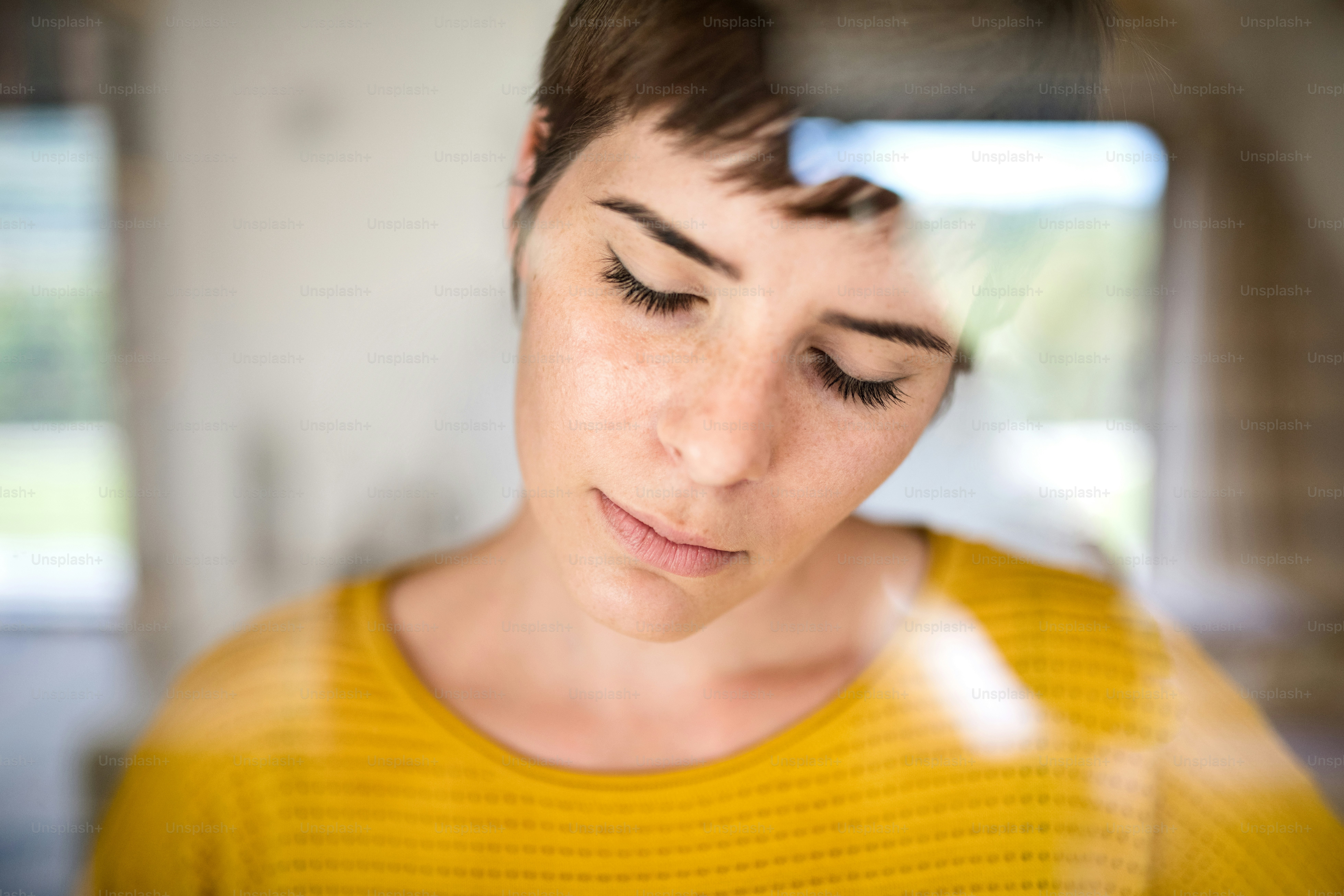 自宅で屋内に立っている悲しい若い女性の正面図、接写。ガラス越しに撮影。