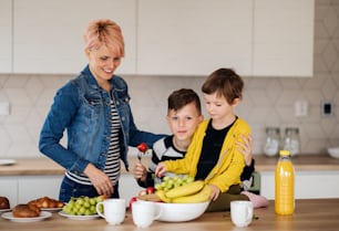 A young woman with two happy children eating fruit in a kitchen.